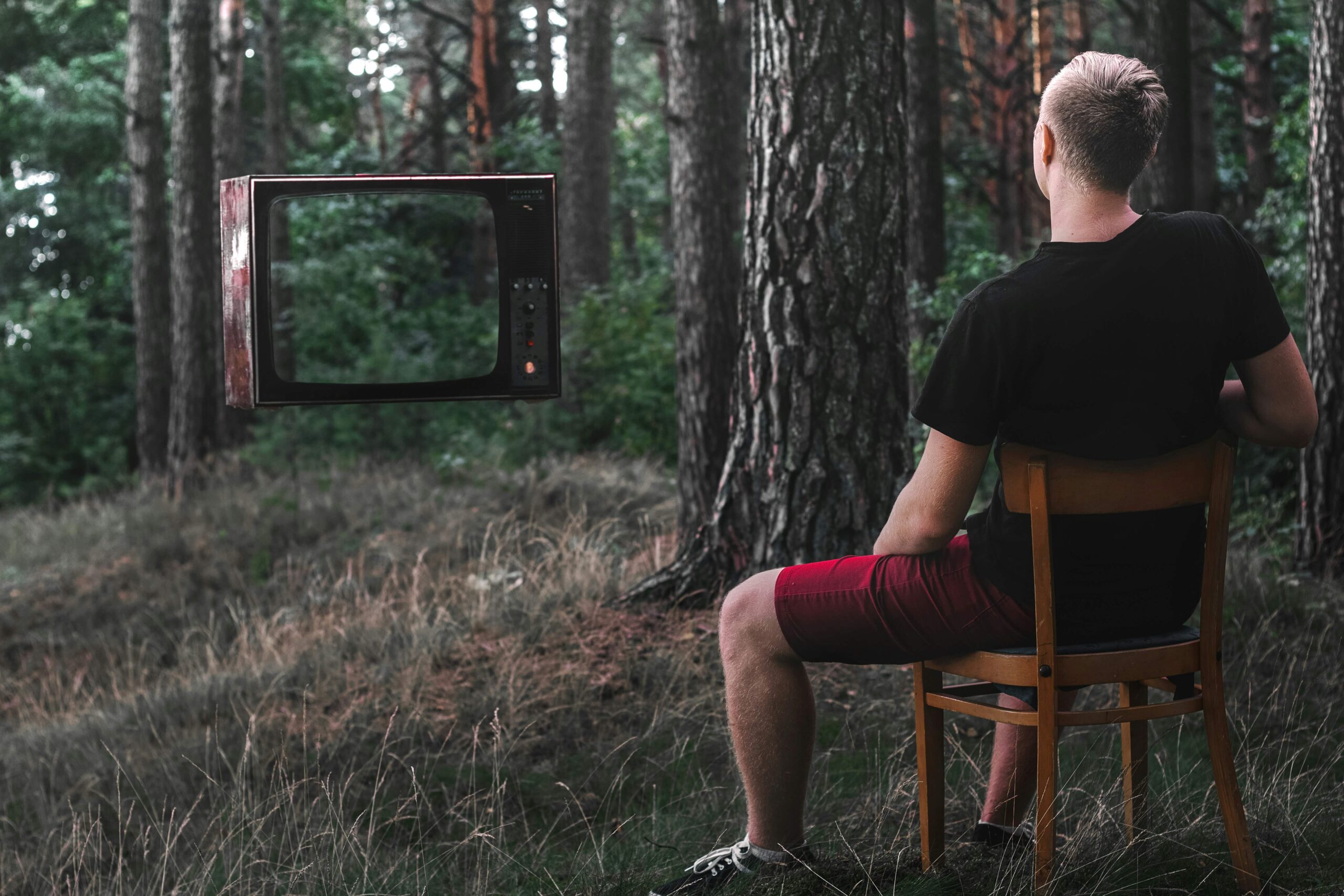 A man sits on a wooden chair in the middle of a forest, gazing through the empty frame of an old television that reveals the trees behind it.