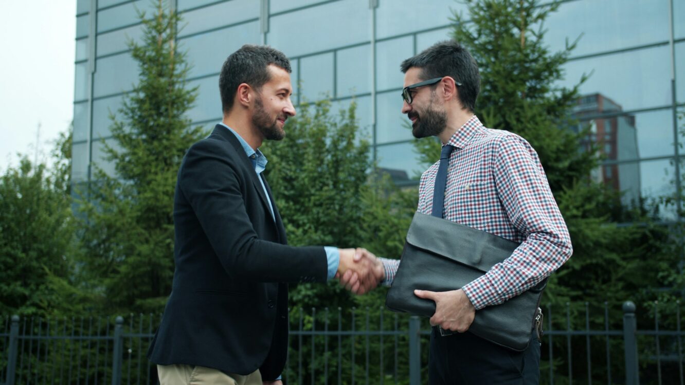 Two business professionals shaking hands outdoors in front of an office building, symbolizing partnership and collaboration for sustainability.